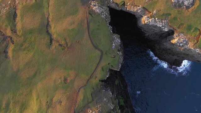 Top aerial view of huge cliff of Faroe Islands, drone flying over a V cove in sunset. Green and brown meadow on stone cliff. Rough blue sea at cliff's feet