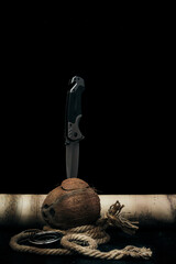Tactical folding knife is stuck in split coconut lying next to rope and an old map on dark...