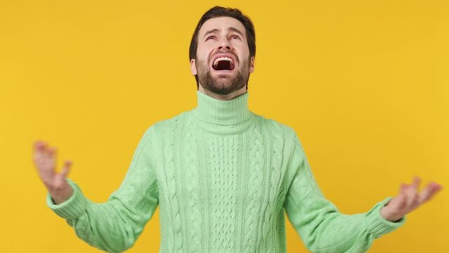 Upset tired young brunet bearded man 20s wears mint shirt looking up screaming crying asking why put hands on face isolated plain yellow background studio portrait. People emotions lifestyle concept
