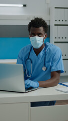 Portrait of man with nurse profession wearing uniform at face mask looking at camera in healthcare office. Young adult sitting at desk while typing on laptop keyboard during pandemic