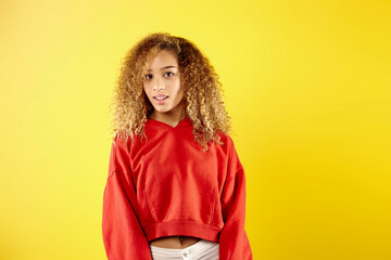 Confident young African American woman in stylish red hoodie and eyeglasses standing on yellow background in studio 