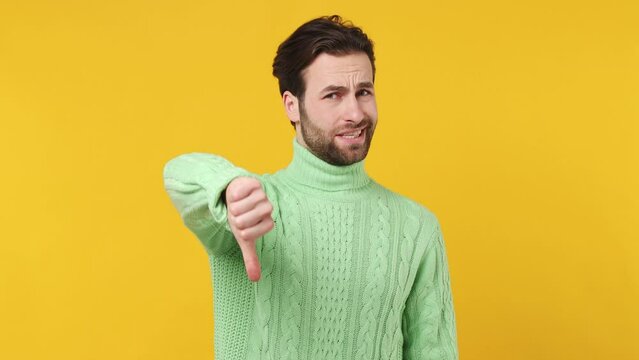 Frowning sad displeased young brunet bearded man 20s years old wears mint shirt showing thumb down dislike gesture isolated plain yellow background studio portrait. People emotions lifestyle concept