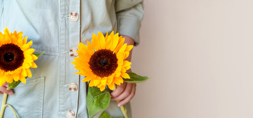 A girl in a light green cotton jumpsuit holds two sunflowers in her hands, a beige monochrome background. The concept of an ecological lifestyle and clothing. copy space