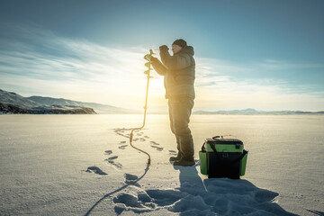 Fisherman drills a hole in the ice of a large frozen lake on a sunny day. The joy of winter fishing