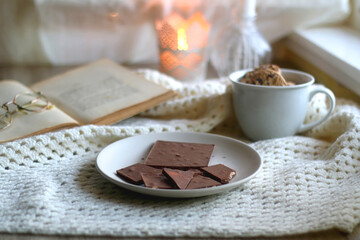 Plate with almond chocolate bar, mug with chocolate chip cookies, open book, reading glasses, soft blanket and lit candles. Hygge at home. Selective focus.
