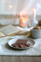 Plate with almond chocolate bar, mug with chocolate chip cookies, open book, reading glasses, soft blanket and lit candles. Hygge at home. Selective focus.