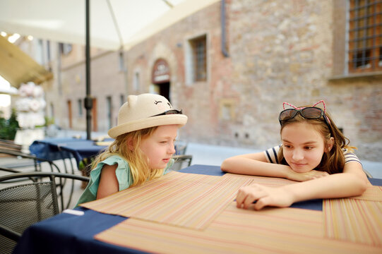 Two Sisters Sitting In Outdoor Pizzeria At Old Narrow Street Of Montepulciano Town, Located On Top Of A Limestone Ridge Surrounded By Vineyards. Tuscany, Italy.