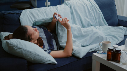 Sick person with fever using oximeter to measure pulse pressure and oxygen saturation while resting on couch with blanket. Woman with disease looking at medical device for diagnosis