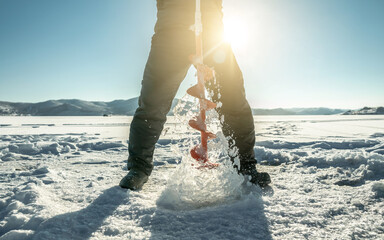 Fisherman drills a hole in the ice of a large frozen lake on a sunny day. The joy of winter fishing