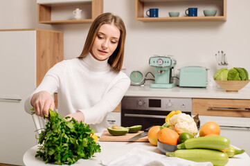 A young, beautiful vegetarian girl or blogger prepares breakfast of fruits, vegetables and greens at home in the kitchen. Blog about healthy eating