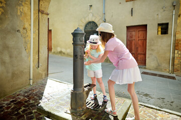Two sisters playing with drinking water fountain in Montalcino town, located on top of a hill top and surrounded by vineyards, known worldwide for the production of wine. Tuscany, Italy.