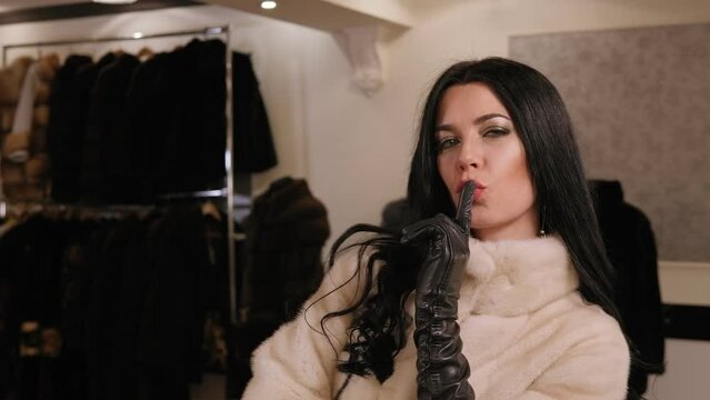 Close-up of a luxurious girl trying on a white fur coat with short arms, leather long gloves in a women's clothing store. Slow motion.