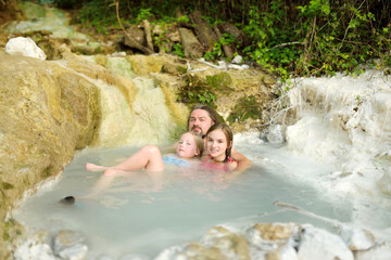 Fototapeta na wymiar Family of three bathing in Bagni San Filippo, small hot spring containing calcium carbonate deposits, forming white concretions and waterfalls. Geothermal pools and hot springs in Tuscany, Italy.