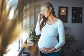 A young Caucasian pregnant woman with a headache is standing by the window in the living room. The...