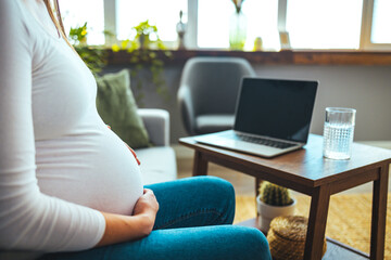 Cheerful mum sitting in chair in bright light flat and using pc laptop while speaking with intelligent doctor about pregnancy. Cropped view of pregnant patient having online consultation with doctor 