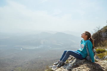 Naklejka na ściany i meble A girl in the mountains sits on a rock and looks at the surrounding landscape. Active sports and recreation.