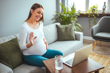 Obraz premium Pregnant woman video chat with family on laptop waving hand at screen. It shows how much the stomach has grown. Young pregnant woman with laptop browsing or chatting with friends online, sitting 