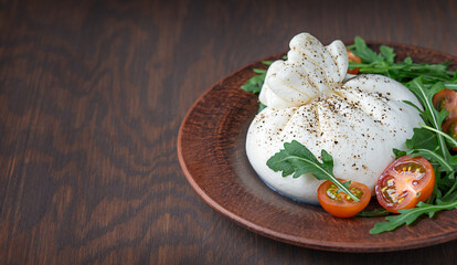 Italian burrata white soft milk lactose cheese made of mozzarella and cream served with sliced red cherry tomatoes and arugula green leaves on plate on dark brown wooden background with copy space
