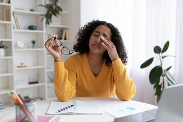 Young black woman feeling tired, rubbing irritated eyes, sitting at desk with laptop, exhausted from studying online - obrazy, fototapety, plakaty