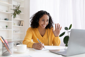 Black woman sitting at table, using laptop, having virtual conference, waving at webcam, greeting...
