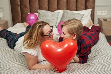 a lesbian couple lies on the bed and smiles at each other, in front of them is a red balloon in the shape of a heart - Powered by Adobe