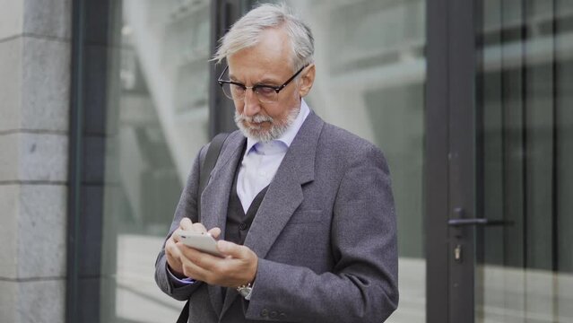 Elegant man in eyeglasses and warm coat standing outdoor looking through news web pages on his smartphone, waiting for friends or dame to go for a walk after work