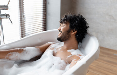 Relaxing bath. Indian man lying in bathtub, enjoying foam at home spa, resting in hot water at...