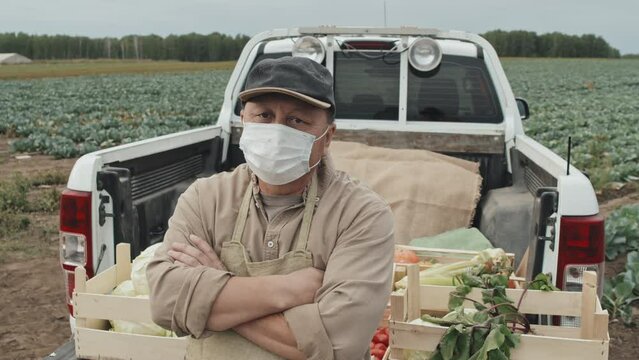 Medium portrait shot footage of unrecognizable mature farm owner wearing mask standing with arms crossed against pickup truck looking at camera