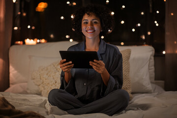technology, bedtime and rest concept - happy smiling woman in pajamas with tablet pc computer sitting in bed at night