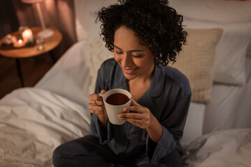 people, bedtime and rest concept - happy smiling woman in pajamas sitting in bed with hot chocolate...