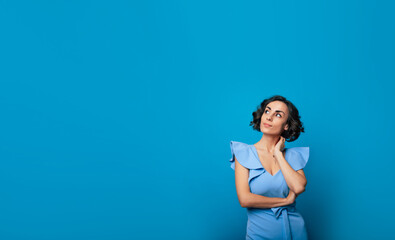 Close-up photo of a charming healthy woman in a blue dress, who is looking in the camera and smiling, while standing and posing isolated on blue background