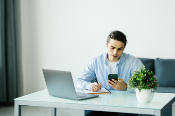 Young happy man smiling while reading his smartphone. Portrait of smiling business man reading message with smartphone in office. Man working at his desk at office.