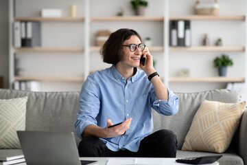 Obraz premium Always in touch. Young freelancer guy talking on cellphone and working on laptop remotely from home, sitting on sofa