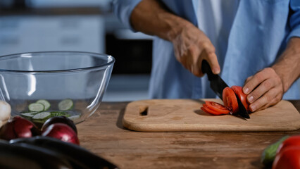 cropped view of man cutting tomato near blurred vegetables and glass bowl.