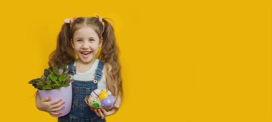 Cheerful little girl with ponytails from her hair holds a basket with Easter eggs and a potted flower in her hands on a yellow background. Copyspace. Layout for advertising. Banner. Horizontal format