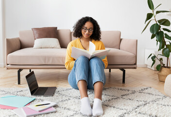 Beautiful young black woman reading book, sitting on floor near laptop pc and study materials at home