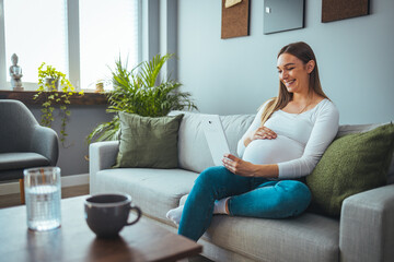 Attractive young pregnant woman smiles while using her digital tablet to video chat while relaxing in her living room. Pregnancy, technology, people and expectation concept