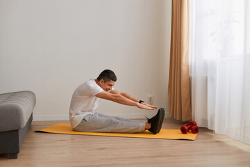 Indoor shot of sporty athletic man wearing sportswear sitting on yoga mat on floor at home and doing stretching, having workout, training flexibility.