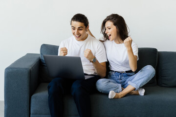 Cheerful loving young couple celebrating internet success, making yes gesture looking at computer...