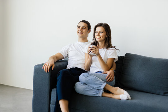 Photo of a young loving couple indoors at home sitting on sofa hugging drinking coffee or tea watch TV.
