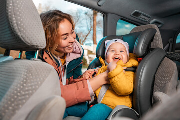 Smiling caucasian mother prepares her little cute child for a trip by car. A woman fastens her seat...