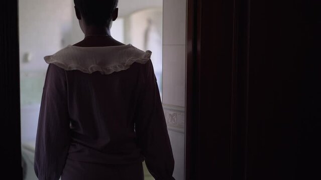 Young slim African American woman in pajamas entering bathroom sitting down on pouf smiling. Portrait of happy relaxed beautiful lady at home indoors in the morning. Slow motion