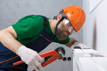 A male plumber installs a radiator in the heating system of an apartment. Guy in overalls and a gas...