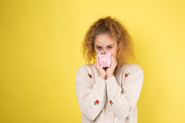 A cute young girl with curly red hair holds a piggy bank, a pink piglet in her hands. The concept...