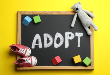 Small chalkboard with word ADOPT, toy bear, colorful cubes and baby shoes on yellow background, top view