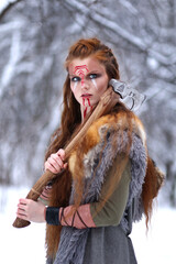 Viking girl with paited face and axe on her shoulder dressed in fur coat in winter forest