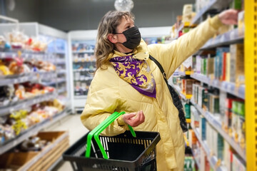 Mature woman in protective mask choosing food products on shelves in grocery shop