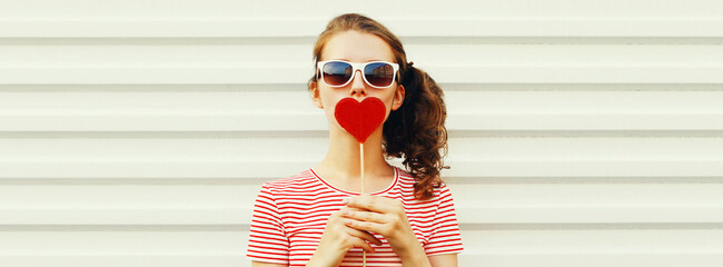 Portrait of beautiful young woman blowing her lips with lipstick with red sweet heart shaped lollipop on white background