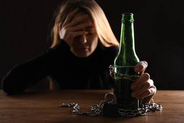Alcohol addiction. Woman chained with bottle of beer at wooden table against black background, focus on hand