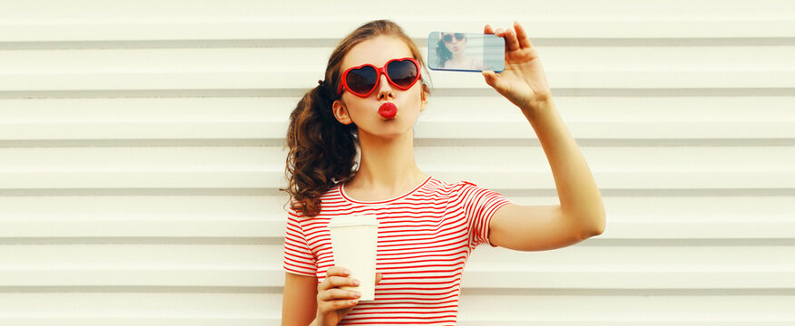 Portrait of young woman taking selfie by phone with coffee cup blowing her lips with red lipstick sending sweet air kiss wearing heart shaped sunglasses on white background